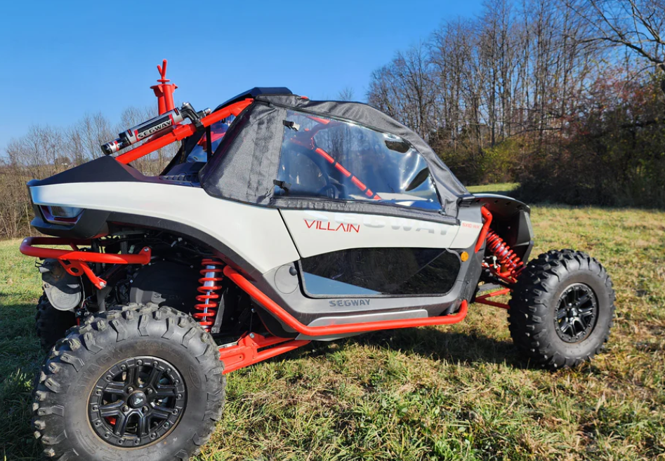 A Segway Villain fitted with the Upper Soft Door/Rear Window Combo, featuring clear plastic windows, is parked on a grassy field with trees and a blue sky in the background. The red and white off-road buggy has rugged tires and exposed suspension.