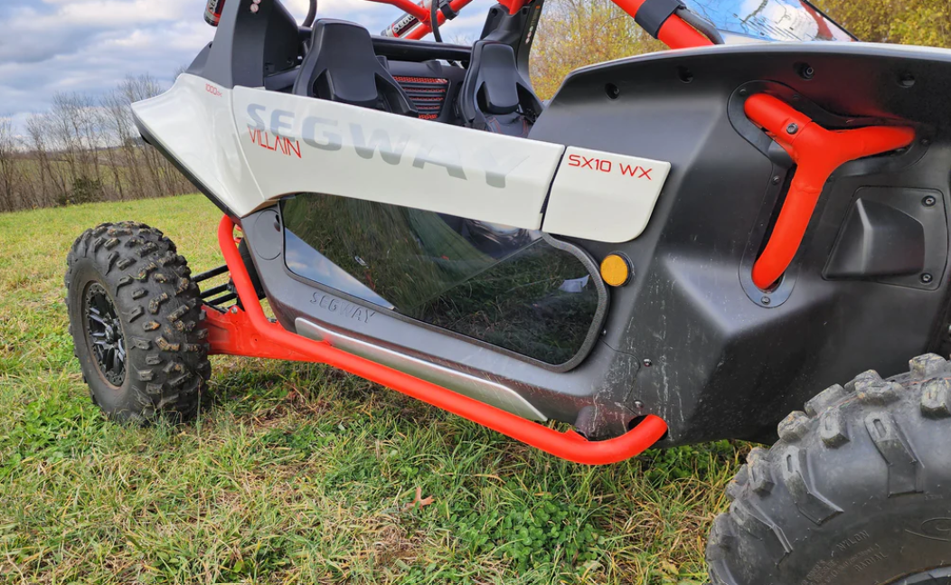 A close-up of Segway Villain with 1/4 polycarbonate lower doors, rugged tires, and red roll cage. The white and black body features bold SEGWAY decals, set on grass with trees in the background.