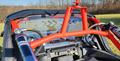 Close-up of an off-road vehicle interior with a bright red roll cage, steering wheel, shock absorbers, and the Segway Villain 3-Piece Polycarbonate Rear Windshield featuring black and yellow arrow decals. Leafless trees and grass are visible outside.