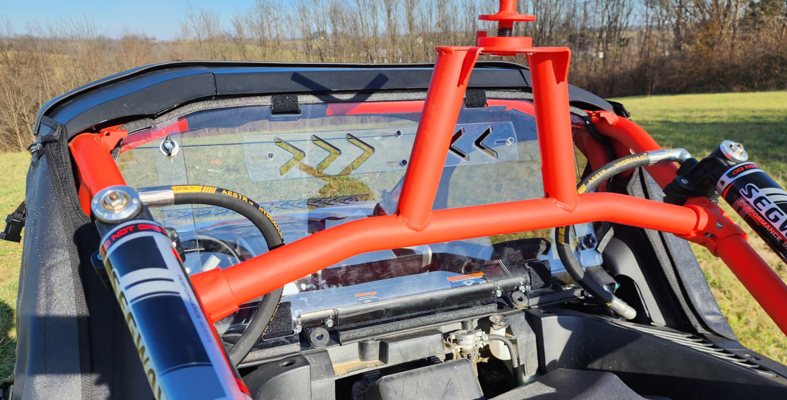 Close-up of an off-road vehicle interior with a bright red roll cage, steering wheel, shock absorbers, and the Segway Villain 3-Piece Polycarbonate Rear Windshield featuring black and yellow arrow decals. Leafless trees and grass are visible outside.