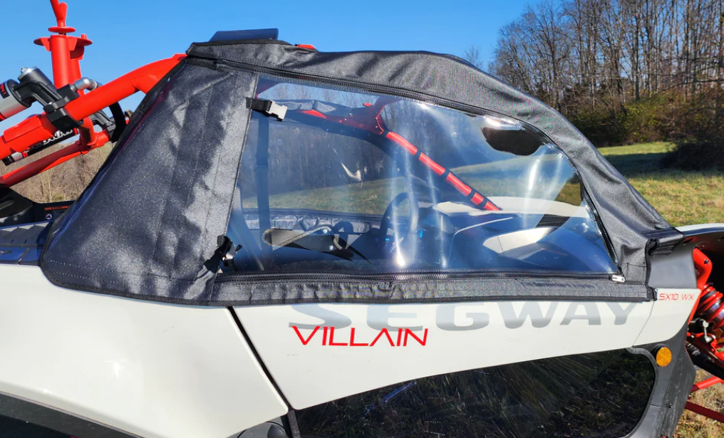 Side view of a Segway Villain with Soft Upper Doors, showing its white body, black soft-top cover, and clear plastic side window. Interior details, red frame accents, bare trees, and a blue sky are visible in the background.