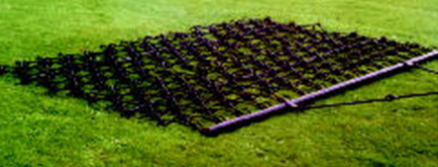 The UTV/ATV 6 Flexible 1/2 Tine Drawbar Harrow, featuring a rectangular grid of spikes, lies flat on green grass. Attached to a bar and chain, its designed to be pulled for agricultural or landscaping tasks.