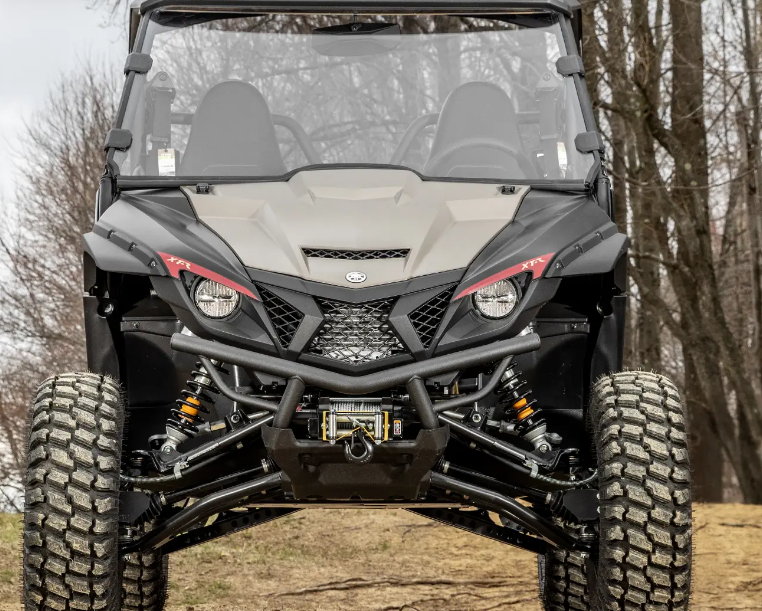 A front view of a black and gray Yamaha Wolverine X2/X4 1000 with a 3 Lift, featuring large rugged tires, angular headlights, and a mesh grille. The vehicle sits on uneven ground with visible suspension beneath the front bumper.