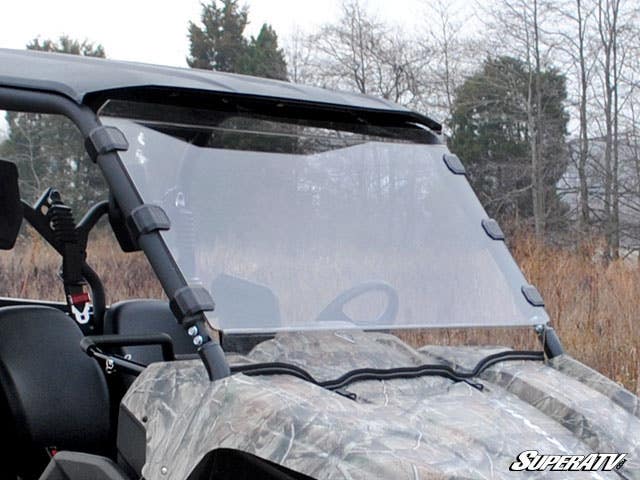 A close-up of a camouflage Yamaha Viking with a slightly tinted Full Windshield made of clear polycarbonate, mounted by black brackets. Leafless trees and dry grass appear in the background as the windshield covers the vehicle’s front.