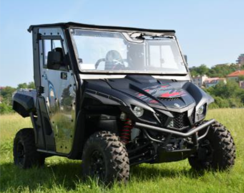 A Yamaha Wolverine 1000 X2 Full Hard Enclosure Cab Kit by Hardcabs, featuring rugged tires, a metal frame, windshield, side doors, and red front accents, is parked on grass with trees and distant buildings under a clear blue sky.