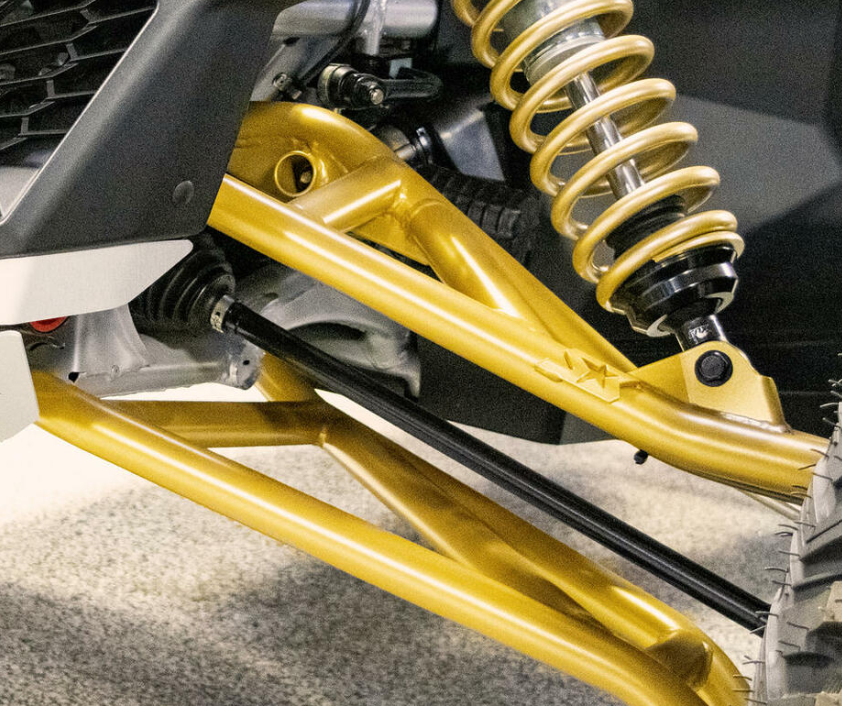 Close-up of the S3 Power Sports 2017+ Can-Am Maverick X3 64 HD High Clearance A-Arm Kit, highlighting its gold-colored suspension with coil spring, shock absorber, and control arms on the wheel hub against a blurred gray floor background.