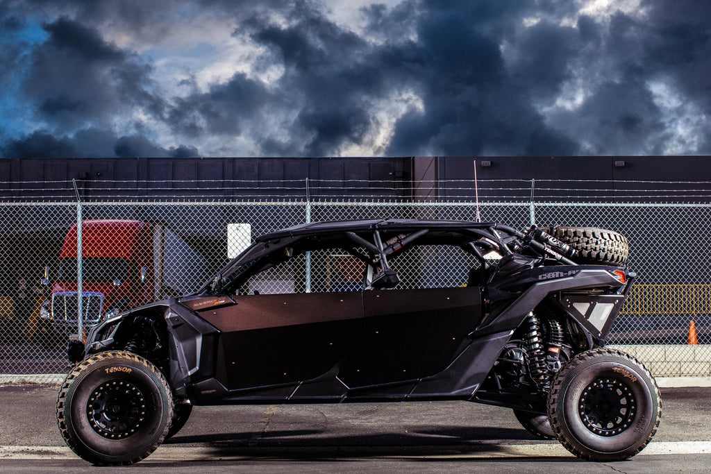 A Canam Maverick X3 Max with Dirt Specialties Aluminum Half Doors is parked by a tall chain-link fence, large rugged tires standing out. Behind it, a red truck cab and industrial building appear under dramatic dusk clouds.