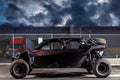A Canam Maverick X3 Max with Dirt Specialties Aluminum Half Doors is parked by a tall chain-link fence, large rugged tires standing out. Behind it, a red truck cab and industrial building appear under dramatic dusk clouds.