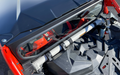 Close-up view of the Maverick X3 Rear Glass Windshield installed on a red off-road vehicle, showing the roll cage, grey support bars, rear glass window, textured black cargo area, and red seats with harnesses visible through the glass.