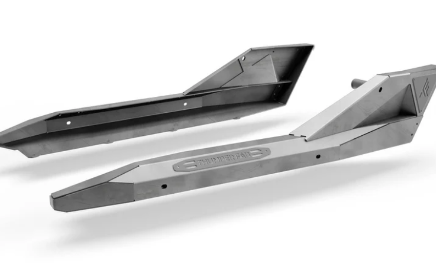 Two metal components—the Maverick X3 Rock Sliders (2 Seat)—with angular, geometric shapes are shown on a white background. One is engraved “THUNDERTRAY”; both have bolt holes and folded edges, indicating they are parts of a larger assembly.