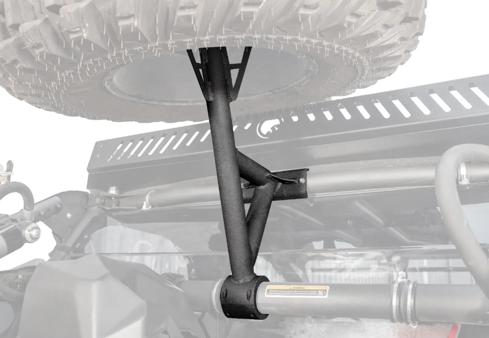 Close-up of the Can Am Maverick X3 Spare Tire Carrier, a rugged black metal mount securely holding a spare tire above the rear cargo area, with visible roll cage, roof rack, and suspension bar; background is faded for emphasis.