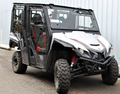 The Yamaha Wolverine X4 Full Hard Enclosure Cab Kit by Hardcabs is shown parked on asphalt next to a corrugated metal wall, featuring four seats with black and red accents, rugged tires, a silver and black body, red shocks, and clear doors.