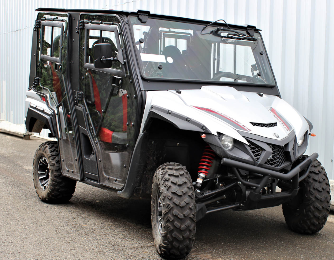 The Yamaha Wolverine X4 Full Hard Enclosure Cab Kit by Hardcabs is shown parked on asphalt next to a corrugated metal wall, featuring four seats with black and red accents, rugged tires, a silver and black body, red shocks, and clear doors.