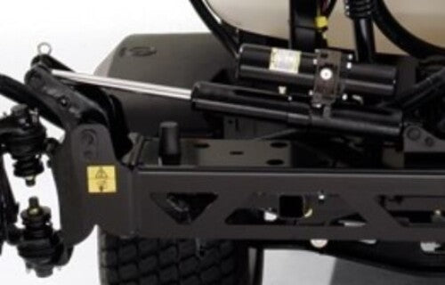 Close-up of the Toro Sprayer Electric Boom Lift Kit with KYB Lift Cylinder Actuator MMP4-B1B250BB-DHR, showing black mechanical suspension, coils, hydraulic tubing, shock absorber, warning sticker on metal parts, and a large tire in the background.