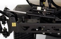 Close-up of the Toro Sprayer Electric Boom Lift Kit with KYB Lift Cylinder Actuator MMP4-B1B250BB-DHR, showing black mechanical suspension, coils, hydraulic tubing, shock absorber, warning sticker on metal parts, and a large tire in the background.