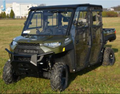 A Polaris Ranger XP1000 Crew with a Hard Cab Enclosure kit, painted green and equipped with off-road tires, is parked on a grassy field. The four-door UTV has clear windows, a rear cargo bed, and buildings and trees in the background.