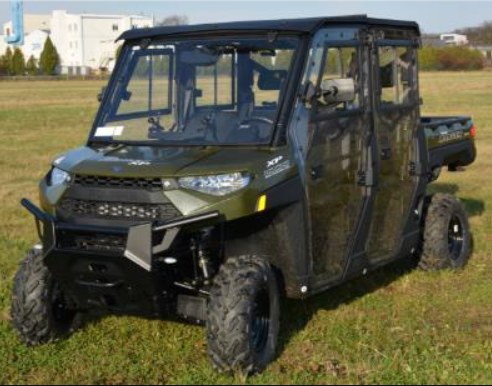 A Polaris Ranger XP1000 Crew with a Hard Cab Enclosure kit, painted green and equipped with off-road tires, is parked on a grassy field. The four-door UTV has clear windows, a rear cargo bed, and buildings and trees in the background.