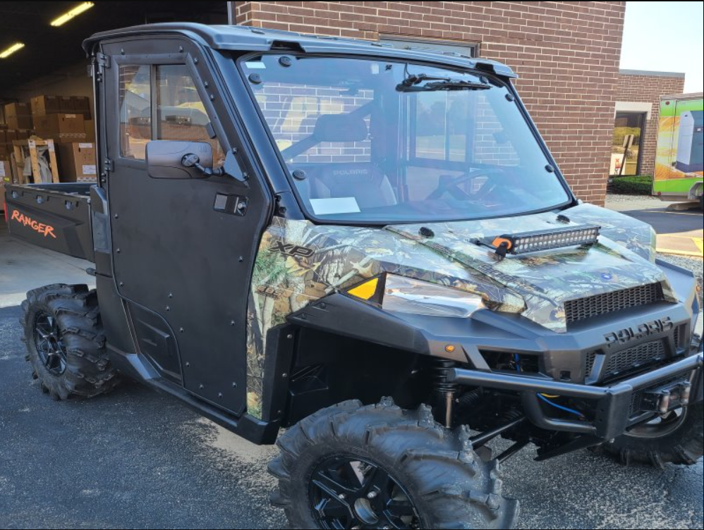 A Polaris Ranger XP900 with the Protector Cab Kit, featuring a camo-patterned hood, black doors, closed cab, cargo bed, and large off-road tires, is parked on pavement outside a brick building.