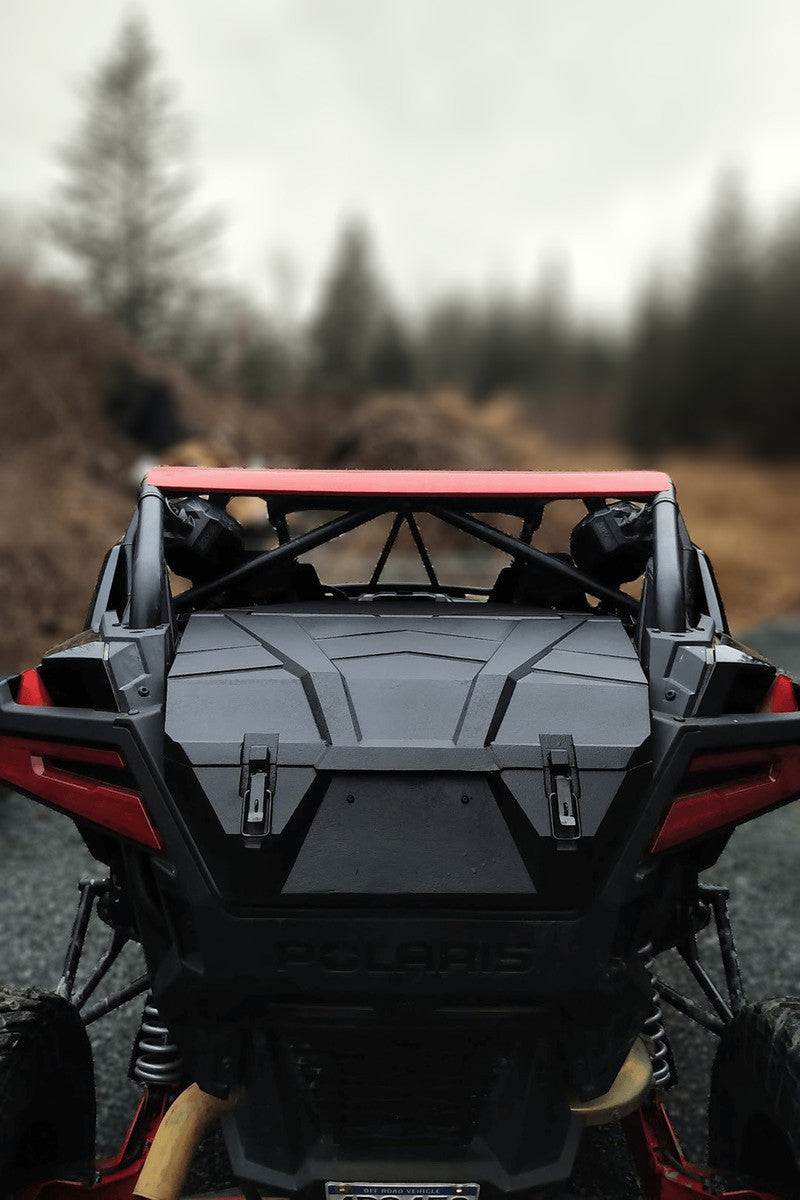 A JEMCO PXP Xtreme UTV Rear Cargo Box is installed on a Polaris RZR Pro XP, parked on gravel among blurred leafless trees and brown brush under an overcast sky. The image highlights the vehicles rugged rear design and suspension.