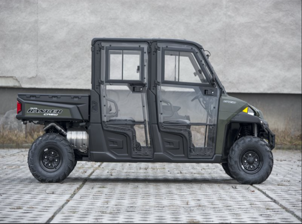 A side view of a green and black Polaris Ranger Crew XP900 Full Hard Cab (2013–PRESENT) utility vehicle parked on pavement, featuring clear hard-sided doors, two-seat rows, four wheels, and a plain light wall in the background.
