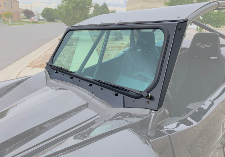 A close-up shows a Wildcat XX Vented Glass Windshield with Wipers on a black off-road vehicle. Through the clear glass, two bucket seats with headrests and logo detailing are visible. The vehicle is parked on a suburban street.