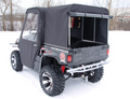 A Yamaha Rhino utility vehicle with off-road tires and a covered cab is parked on snow. It features a Yamaha Rhino Soft Bed Cover Shell with a window at the back, surrounded by snowy trees and a fence in the background.