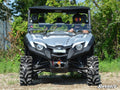 A gray Yamaha Viking equipped with a 2 Lift Kit is parked on gravel, facing forward. A helmeted driver sits at the wheel. Its large tires and front winch are visible, with green trees and bushes in the background.