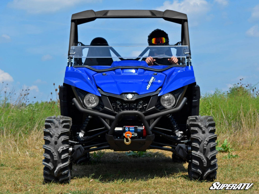 A Yamaha Wolverine 2 Lift Kit-equipped ATV with large, rugged tires and a visible front winch is parked on grass. A goggle-wearing driver sits at the wheel, surrounded by wildflowers and a bright blue sky.