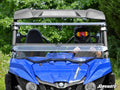 A person in a black helmet and goggles drives a blue Yamaha Wolverine equipped with a Yamaha Wolverine Flip Windshield. Green trees are in the background, and the SuperATV logo appears in the lower right corner of the image.