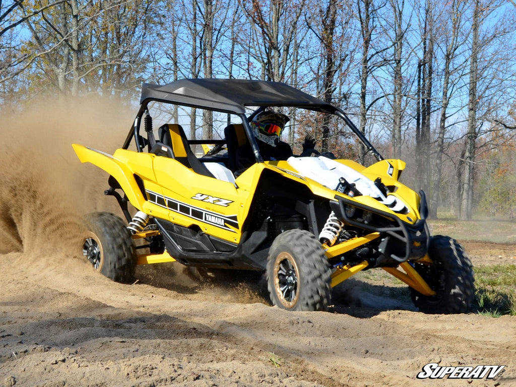 A Yamaha YXZ equipped with a Performance Flywheel tears around a rugged trail, kicking up sand as the helmeted driver powers through a bend. Leafless trees and blue sky frame the action, with the SuperATV logo in the corner.