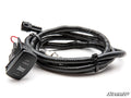 The image shows a Yamaha Wolverine RMAX Ready-Fit Winch harness: a coiled black wiring assembly with a rocker switch labeled WINCH IN/OUT, connector plug, and black corrugated tubing. White background with SuperATV branding in the corner.