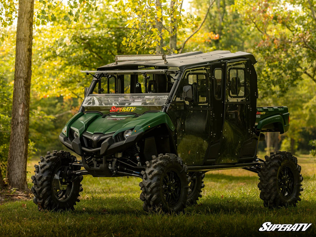 A Yamaha Viking with a 6 Portal Gear Lift and large tires is parked on grass in a sunlit wooded area. It features a roof, doors, and SuperATV logos on the windshield and corner. Trees with green foliage are in the background.