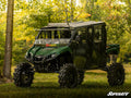 A Yamaha Viking with a 6 Portal Gear Lift and large tires is parked on grass in a sunlit wooded area. It features a roof, doors, and SuperATV logos on the windshield and corner. Trees with green foliage are in the background.
