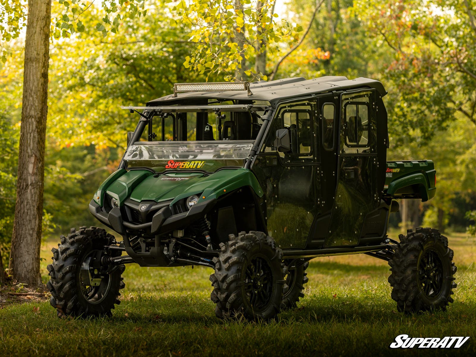 A Yamaha Viking with a 6 Portal Gear Lift and large tires is parked on grass in a sunlit wooded area. It features a roof, doors, and SuperATV logos on the windshield and corner. Trees with green foliage are in the background.