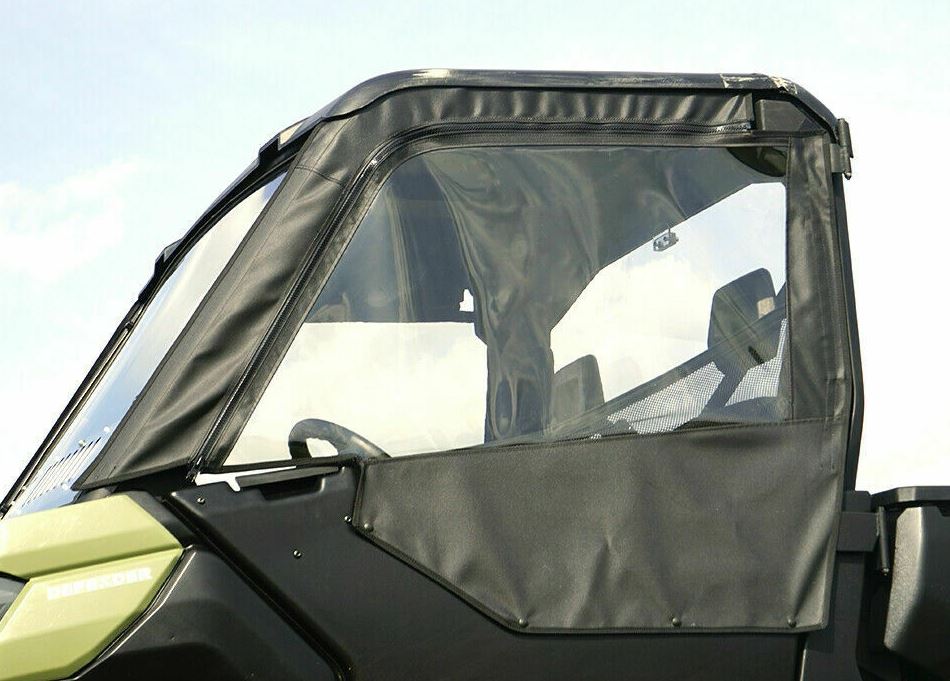 A close-up shows a Canam Defender Upper Soft Door (fits all brand lower doors) with a clear vinyl window panel and visible stitching, covering the side and door. The black enclosure has a slightly shiny finish, with the sky in the background.
