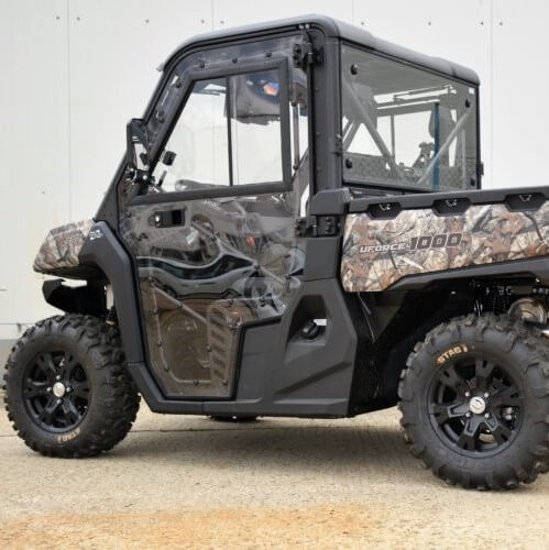 A side view of the Cf Moto Uforce 1000 Full Cab Enclosure UTV shows a rugged vehicle with camo patterns, black wheels, off-road tires, glass doors and windows, and a cargo bed labeled 1000, set against a plain light-colored wall.